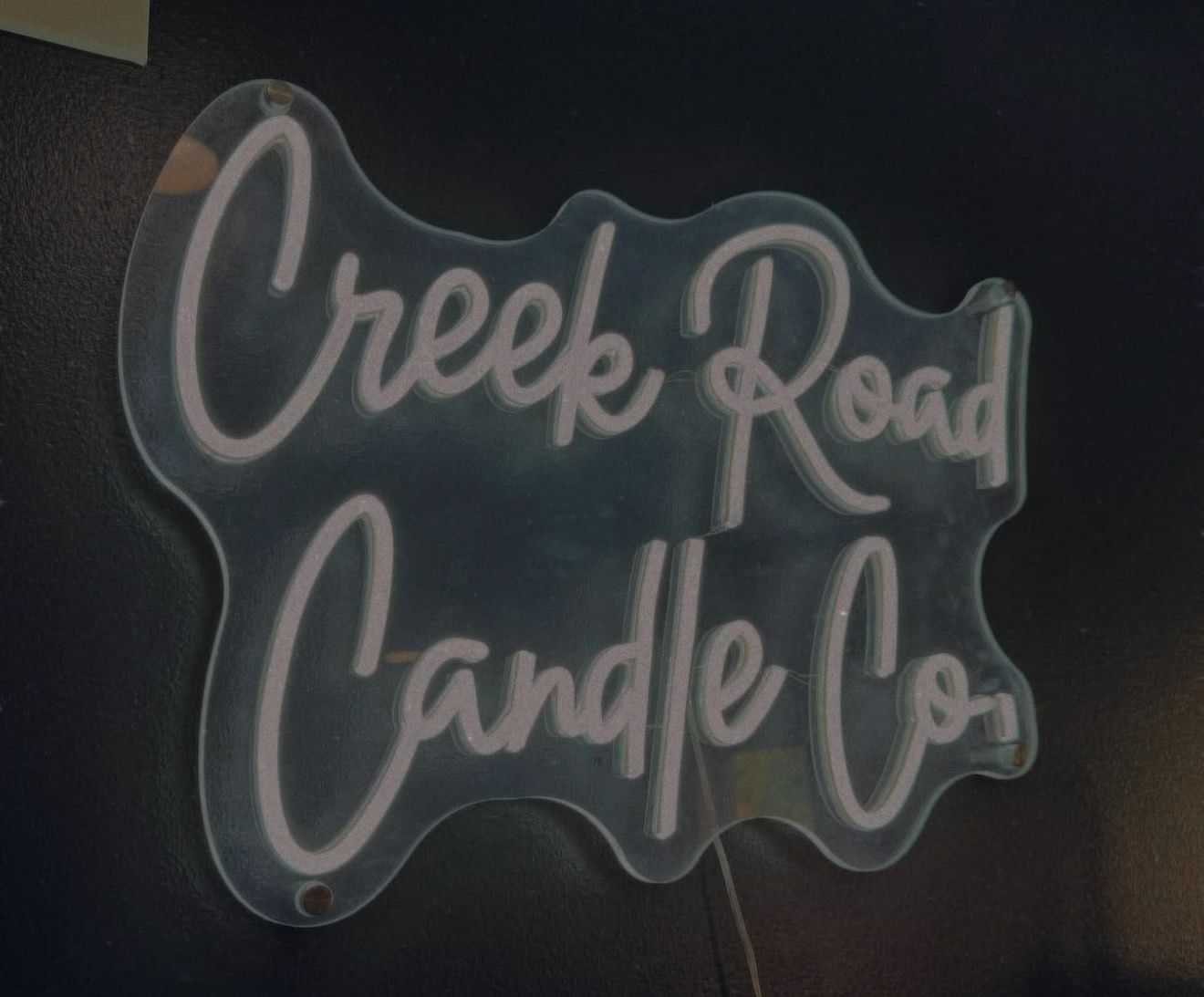 Sign reading "Creek Road Candle Co." in white script on a wavy clear background against a dark wall.