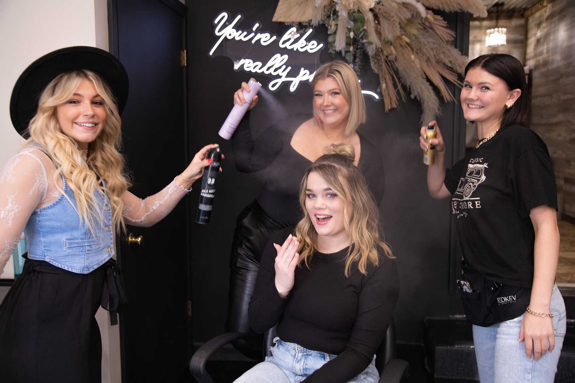 Four women smiling and spraying hair products in a modern salon with a neon sign in the background.