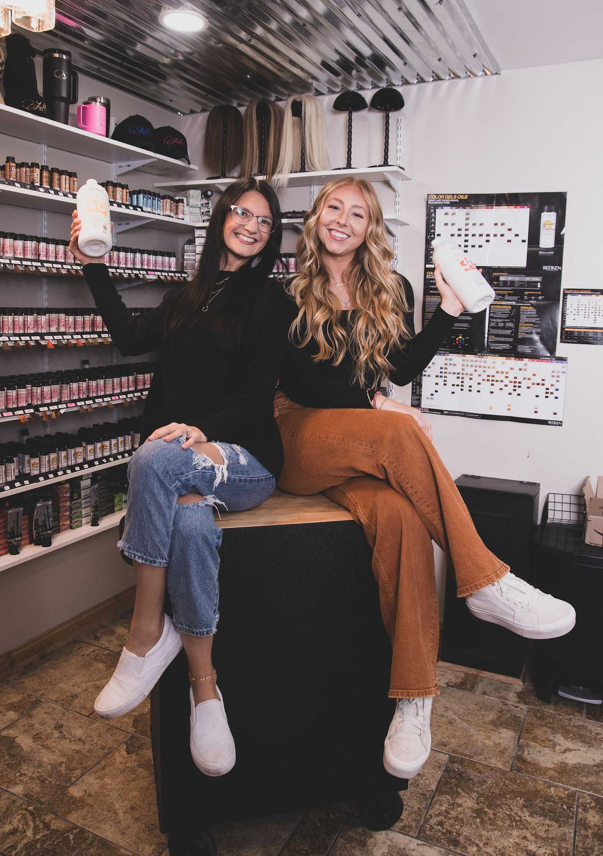 Two smiling women holding hair product bottles in a beauty supply store surrounded by shelves.